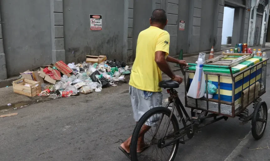 Homem conduz triciclo de carga por uma rua, ao lado de resíduos descartados junto a um muro.
